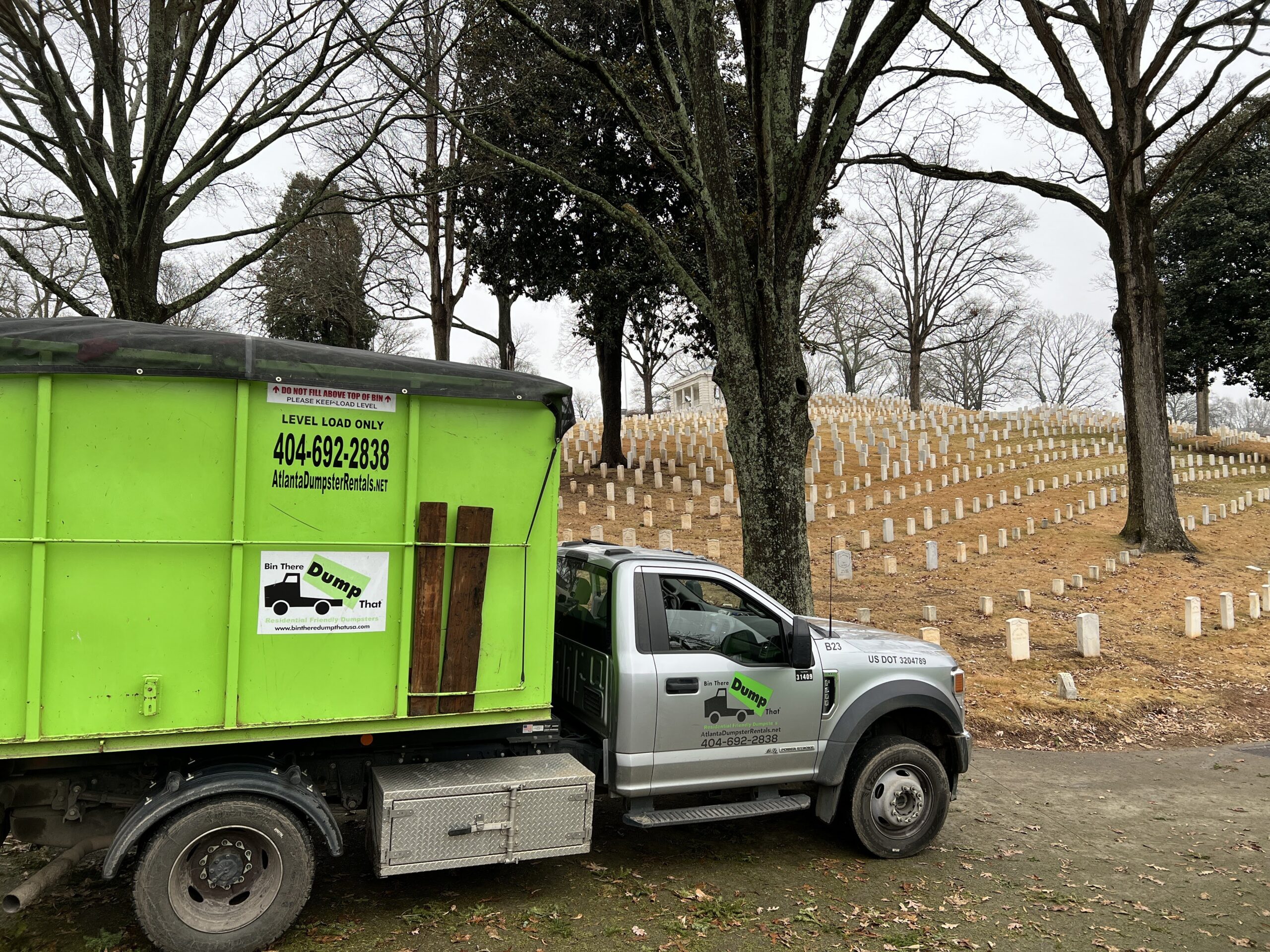 A loaded green Bin There Dump That dumpster on a hook lift truck at Marietta National Cemetery, with rows of white headstones rising on the hillside behind it.