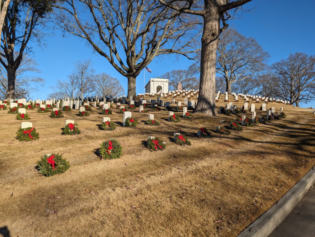 Rows of white headstones on a hillside at Marietta National Cemetery, each with a balsam wreath and red bow at its base, with the stone rostrum and flag at the crest.