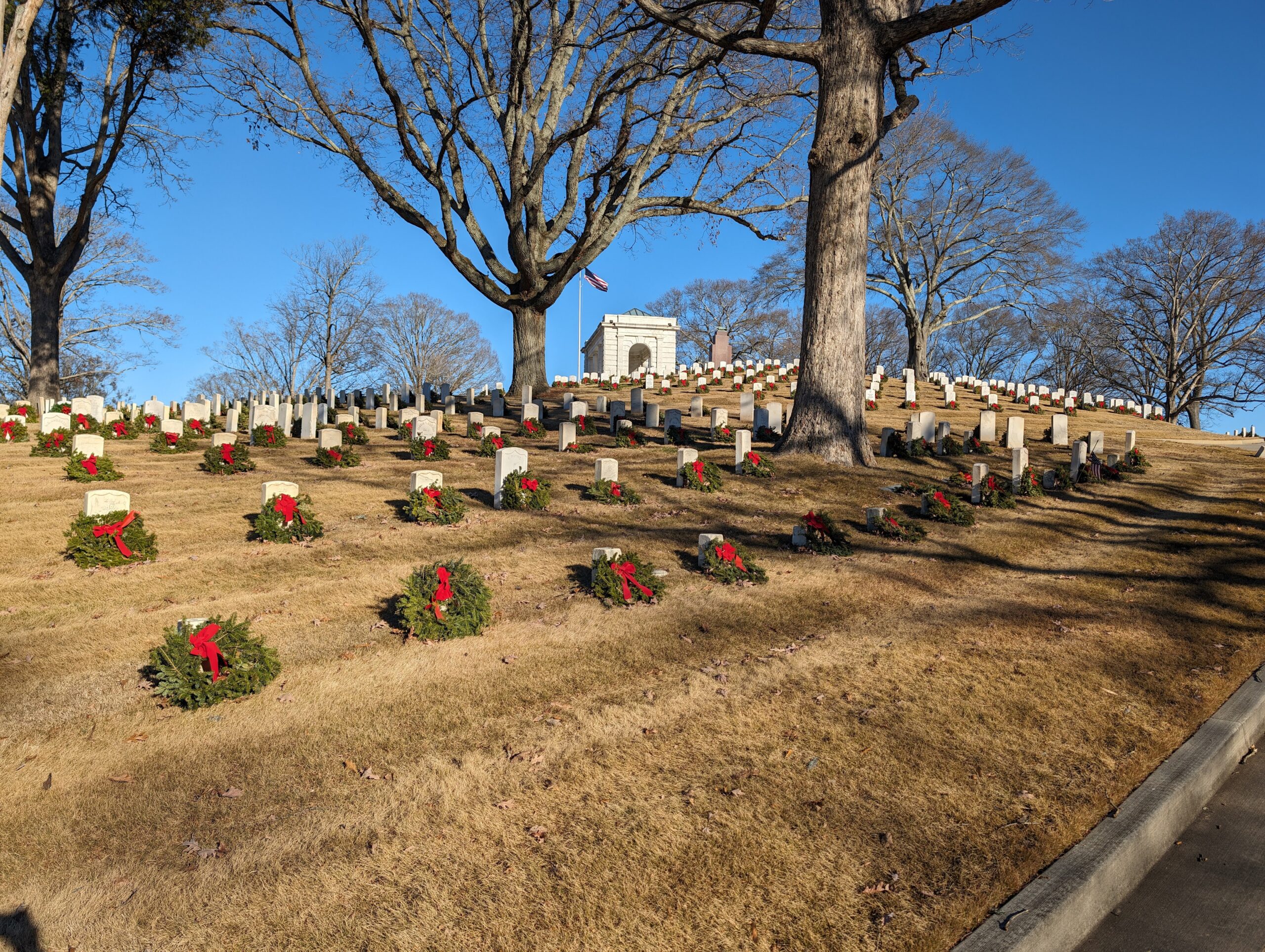 Rows of white headstones on a hillside at Marietta National Cemetery, each with a balsam wreath and red bow at its base, with the stone rostrum and flag at the crest.